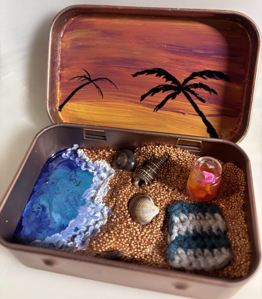 A small container that shows a beach scene. On the lid is a purple and yellow sky with black palm tree outlines and inside the container there is a painted sea and coppery sand with seashells, a fruity drink and a little crochet towel.